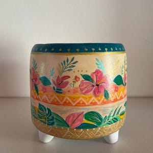 Peut inclure: Un cache-pot cylindrique en céramique sur trois petits pieds blancs, orné de motifs peints à la main de fleurs d'hibiscus roses, de feuilles tropicales vertes et de formes géométriques orange sur fond crème, avec un bord bleu sarcelle.