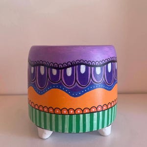 Op de afbeelding: Handbeschilderde, cilindrische bloempot met een levendig ontwerp. De bovenkant is paars met een geschulpte zwarte rand, gevolgd door oranje, blauwe en groen gestreepte secties. Hij staat op drie witte voetjes, perfect voor kleine planten.
