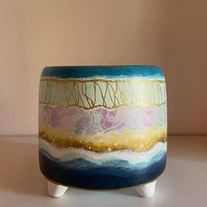 May include: A cylindrical ceramic planter with three small white feet, featuring horizontal bands of colour including deep navy blue, light blue, soft pink, and yellow, accented with gold metallic lines and small white dots throughout the design.