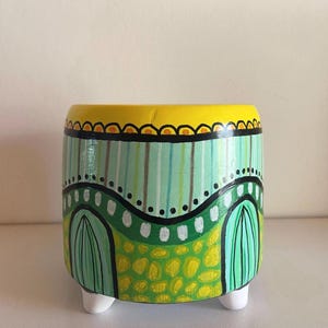 Op de afbeelding: Een handbeschilderde keramische plantenbak met een gele rand en witte voetjes. De body heeft een groen en geel landschapsontwerp met verticale strepen en boogvormen. De plantenbak is een decoratief item.