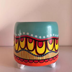 Peut inclure: Un pot de fleurs en céramique peint à la main, avec un dessus turquoise et un motif vibrant. Le motif présente des accents jaunes, rouges et oranges avec des points et des rayures blancs. Le pot repose sur trois petits pieds blancs.