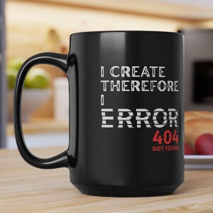 Peut inclure: Mug en céramique noire avec une grande anse. Le mug affiche le texte "I CREATE THEREFORE I ERROR 404 NOT FOUND" en blanc et rouge. Le mug est posé sur une surface en bois.