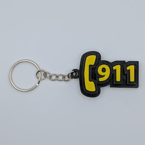 May include: Black and yellow 911 keychain. The keychain features the number 911 in yellow, outlined in black, with a yellow phone receiver shape. The keychain is attached to a silver key ring and chain.