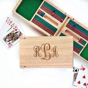 May include: A wooden cribbage board with a personalized monogram, cards, and pegs. The board is open, revealing green felt and red and green scoring tracks. Playing cards are scattered around the board.