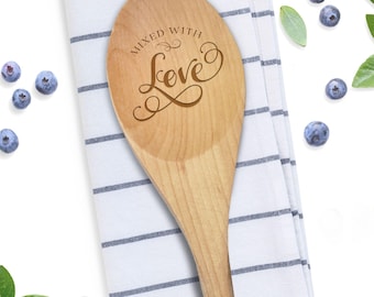 May include: A light-colored wooden spoon featuring the engraved text "MIXED WITH Love" in a decorative script font. The spoon is positioned on a white cloth napkin with thin horizontal blue stripes, surrounded by scattered fresh blueberries.