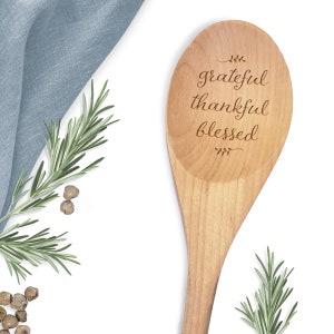 May include: Wooden cooking spoon with the words "grateful, thankful, blessed" engraved on the surface. The spoon is surrounded by sprigs of rosemary and peppercorns, with a blue linen napkin in the background.