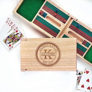 May include: A wooden cribbage board game set with a personalized wooden box. The box has a circular design with the text "Jackson & Haley Kilgore Est. 2022".