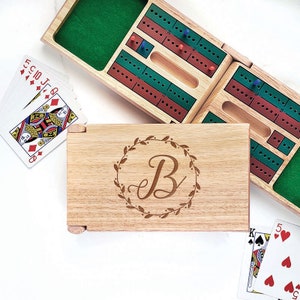 May include: A wooden cribbage board with a green felt interior and red and green scoring tracks. The board is open, revealing the peg holes. A wooden box with a floral wreath and the letter "B" is in the foreground. Playing cards are scattered around.