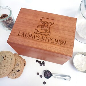 May include: A wooden box with a lid, engraved with a kitchen mixer and the text "LAURA'S KITCHEN". The box is on a white surface with chocolate chips, cookies, and a measuring spoon.