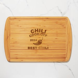May include: A rectangular bamboo cutting board with rounded edges. The board features engraved text that reads "CHILI COOK-OFF 2024 BEST CHILI" along with a chili pepper graphic. The board has a juice groove around the perimeter.