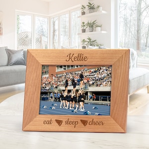 May include: A wooden picture frame with a photo of a cheerleading squad performing on a blue track. The frame is engraved with the name "Kellie" at the top and the words "eat, sleep, cheer" at the bottom. The frame is made of light brown wood.