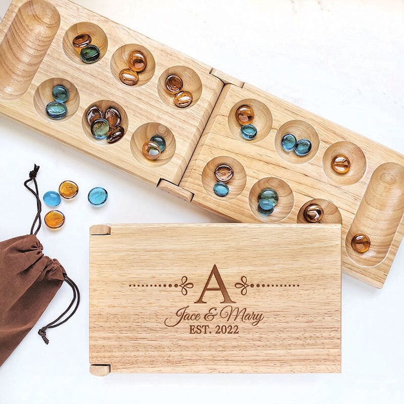 May include: A wooden Mancala board game with a personalized wooden box. The box is engraved with the letter "A", "Jace & Mary", and "EST. 2022". The board game has two rows of six holes, and the holes are filled with brown and blue glass marbles.