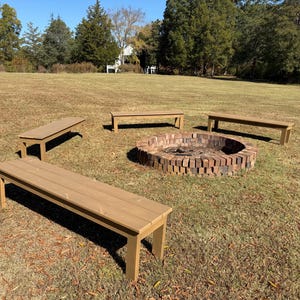May include: An outdoor fire pit surrounded by four wooden benches. The fire pit is made of brick and is in the center of the benches. The benches are made of wood and are a light brown color. The scene is set on a grassy lawn.