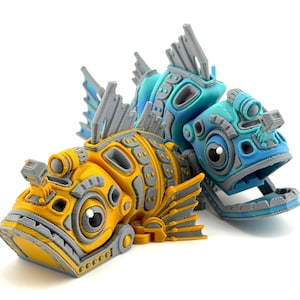 May include: Two 3D-printed fish sculptures. One is yellow and gray, the other is blue and gray. Both have detailed mechanical designs, including fins, eyes, and articulated jaws. The sculptures are set against a white background.