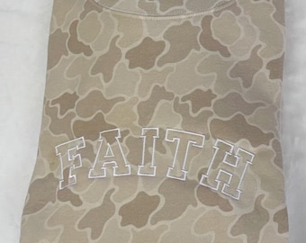 May include: A beige camouflage-patterned sweatshirt with the word "FAITH" embroidered in white block letters across the chest. The sweatshirt has a crew neck and long sleeves. The fabric has a soft, muted color palette.
