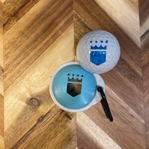 May include: A white golf ball and a light blue ball marker with a crown design. The golf ball has a blue crown design. A black clip is attached to the ball marker. The items are on a wooden surface.