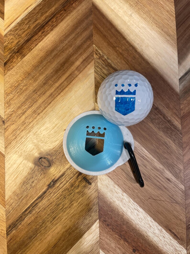 May include: A white golf ball and a light blue ball marker with a crown design. The golf ball has a blue crown design. A black clip is attached to the ball marker. The items are on a wooden surface.