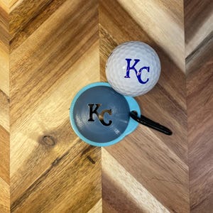 May include: A white golf ball with the letters "KC" in blue, resting next to a blue and gray disc-shaped item with the same initials. The disc has a black loop attached. The items are on a wooden surface.