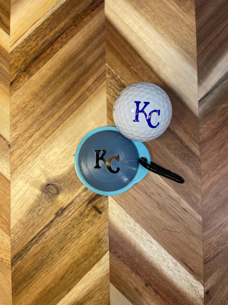 May include: A white golf ball with the letters "KC" in blue, resting next to a blue and gray disc-shaped item with the same initials. The disc has a black loop attached. The items are on a wooden surface.