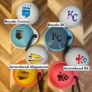 May include: Golf balls with various designs and matching ball markers. The golf balls feature designs such as a crown, the letters "KC", and an arrowhead. The ball markers are in blue, yellow, and pink with a black carabiner clip.
