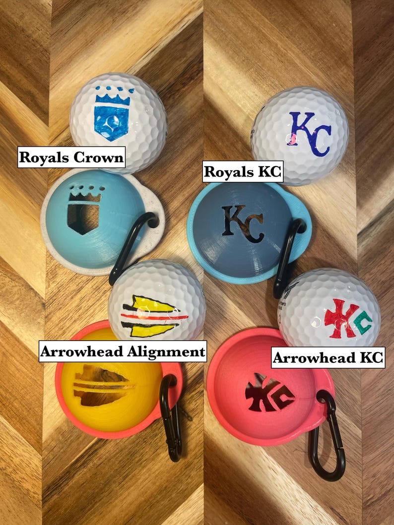 May include: Golf balls with various designs and matching ball markers. The golf balls feature designs such as a crown, the letters "KC", and an arrowhead. The ball markers are in blue, yellow, and pink with a black carabiner clip.