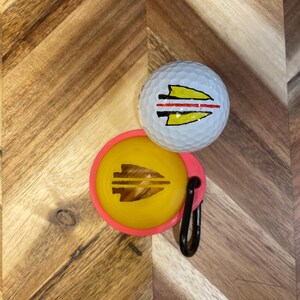 May include: A white golf ball and a yellow ball marker with a pink border. Both items feature a yellow and black arrowhead design. A black carabiner clip is attached to the ball marker. The items are on a wooden surface.