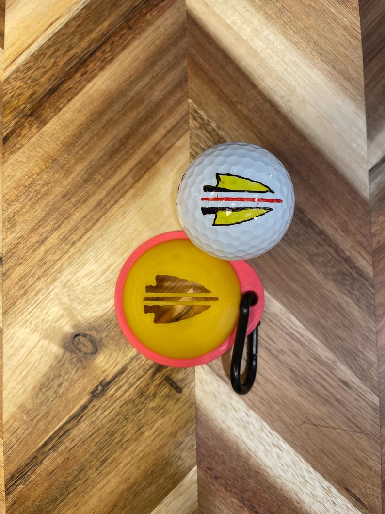 May include: A white golf ball and a yellow ball marker with a pink border. Both items feature a yellow and black arrowhead design. A black carabiner clip is attached to the ball marker. The items are on a wooden surface.