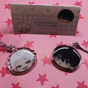 Puede incluir: Dos colgantes para teléfono de estilo anime con ilustraciones de personajes. Uno tiene cabello blanco, el otro cabello negro. Los colgantes están sobre una superficie rosa con forma de estrella. Una caja con el texto "Yoshiki & Hikaru Phone Charms" está en el fondo.