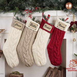 May include: Four cable-knit Christmas stockings in cream, beige, and red, each with a personalized name tag. The names are Claire, Bella, Violet, and Nancy. The stockings are displayed on a fireplace mantel decorated with greenery and ornaments.