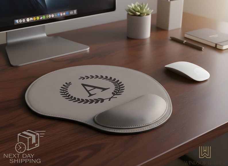 K&ouml;nnte beinhalten: Ein graues Leder-Mauspad mit Handgelenkauflage, mit einem schwarzen Lorbeerkranz-Design und dem Buchstaben "A". Das Pad liegt auf einem Holzschreibtisch, neben einer wei&szlig;en Maus und einer kleinen Topfpflanze.