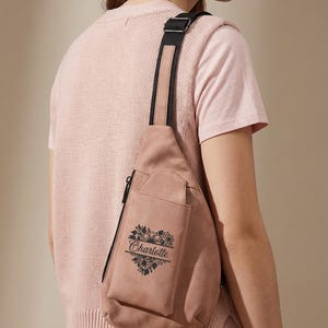 May include: A dusty rose sling bag with a black strap and zipper. The bag features a floral heart design with the name "Charlotte" printed in black. The bag is made of suede-like material.