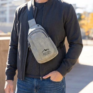 May include: A gray sling bag with a zippered pocket and adjustable strap. The bag features the text "BRIAN TAYLOR" with a mountain logo. The bag is worn across the body.