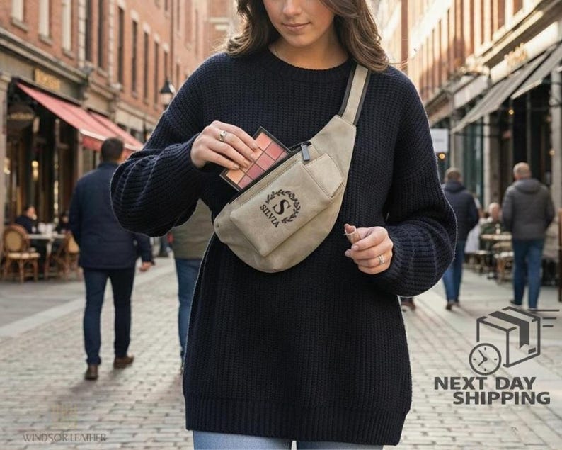 May include: A beige fanny pack with a black strap and zipper, worn across the body. The pack has the name "SILVIA" and a decorative "S" initial. The person is holding a makeup palette. The background shows a city street.