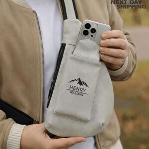 May include: A light gray crossbody bag with a black zipper and a pocket. The pocket has a mountain graphic and the text "HENRY WILLIAMS". A smartphone is in the bag's pocket. The bag has a black strap.