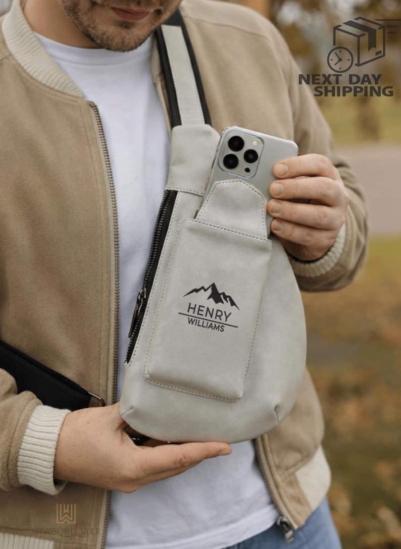 May include: A light gray crossbody bag with a black zipper and a pocket. The pocket has a mountain graphic and the text "HENRY WILLIAMS". A smartphone is in the bag's pocket. The bag has a black strap.