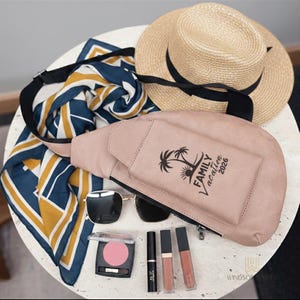 May include: A collection of travel accessories including a light pink fanny pack with "Family Vacation 2026" text, sunglasses, a straw hat with a black band, a patterned scarf, and makeup.