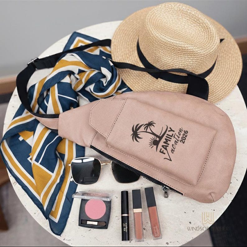 May include: A collection of travel accessories including a light pink fanny pack with "Family Vacation 2026" text, sunglasses, a straw hat with a black band, a patterned scarf, and makeup.