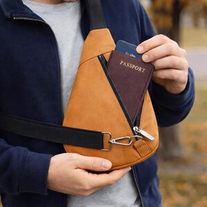 May include: A tan sling bag with a black strap and silver hardware. The bag has a zippered pocket and a passport is visible inside. The person is wearing a navy blue jacket and a gray shirt.