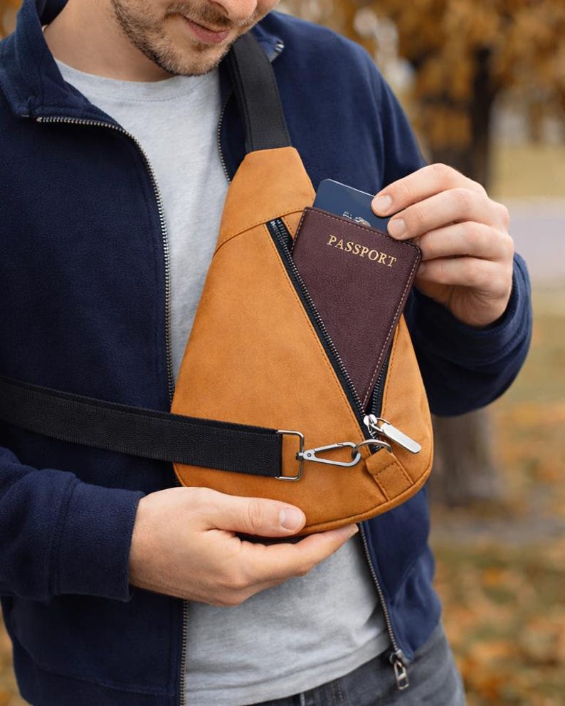 May include: A tan sling bag with a black strap and silver hardware. The bag has a zippered pocket and a passport is visible inside. The person is wearing a navy blue jacket and a gray shirt.