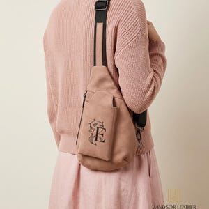 May include: A dusty rose-colored sling bag with a black strap. The bag features a front pocket with a decorative black embroidered design. The bag is worn over the shoulder. The brand name "WINDSOR LEATHER" is visible.