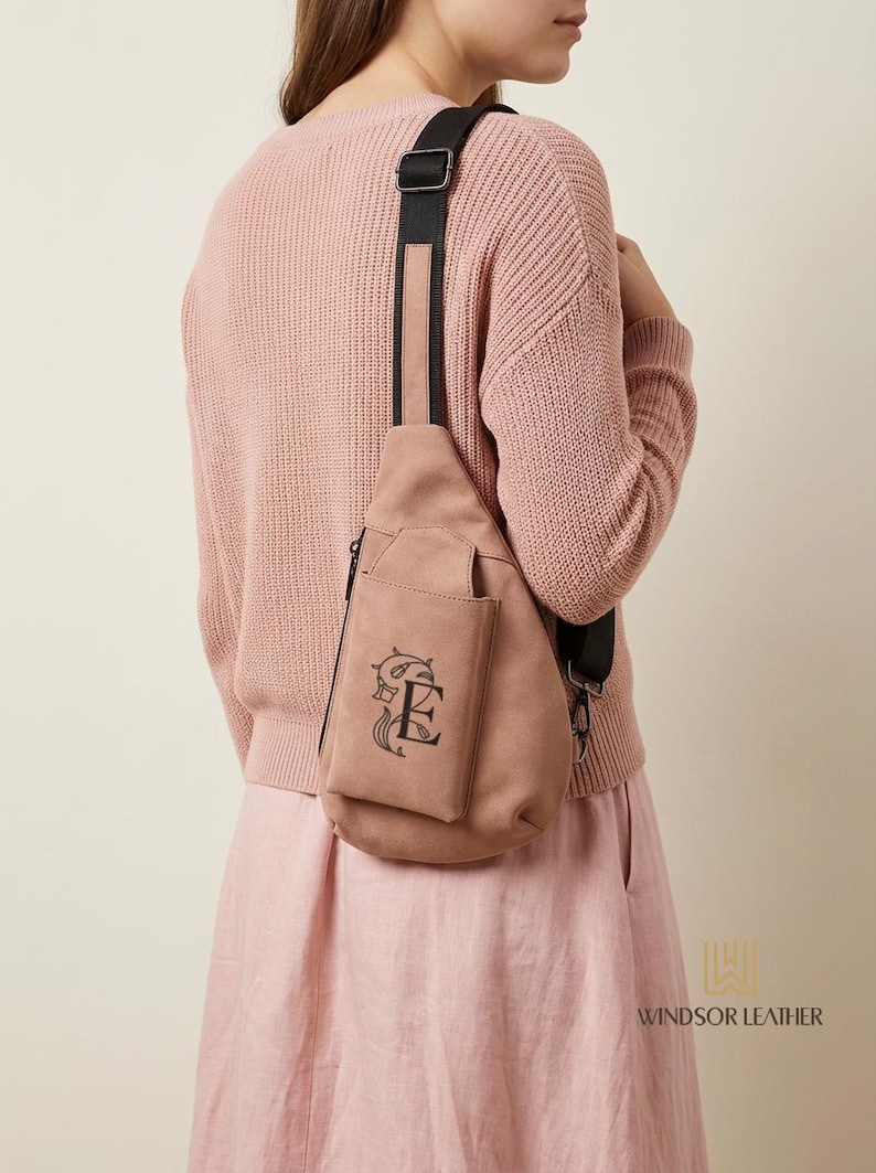May include: A dusty rose-colored sling bag with a black strap. The bag features a front pocket with a decorative black embroidered design. The bag is worn over the shoulder. The brand name "WINDSOR LEATHER" is visible.