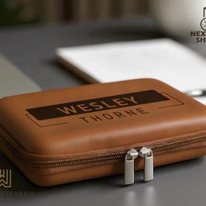 May include: A brown leather case with a zipper closure and silver-colored zipper pulls. The case has the name "WESLEY THORNE" printed on the top. The case is sitting on a gray surface, with a laptop and notepad in the background.