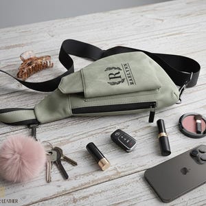 May include: A sage green crossbody bag with a black strap, a zipper, and the name "RAELYNN" printed on the front. The bag is surrounded by keys, a car key, a phone, makeup, and a pink pom-pom.