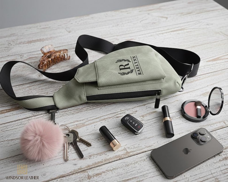 May include: A sage green crossbody bag with a black strap, a zipper, and the name "RAELYNN" printed on the front. The bag is surrounded by keys, a car key, a phone, makeup, and a pink pom-pom.