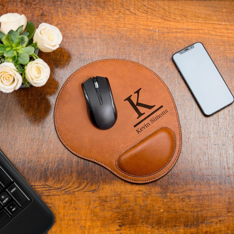 K&ouml;nnte beinhalten: Ein braunes Leder-Mauspad mit einer schwarzen Maus, einem Telefon und einer Laptop-Tastatur auf einer Holzoberfl&auml;che. Das Mauspad hat eine eingebaute Handballenauflage und ist mit dem Buchstaben "K" und dem Namen "Kevin Simons" personalisiert.