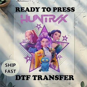 May include: A DTF transfer featuring three anime-style women in front of a purple starburst design. The word "HUNTRAX" is above the women. The words "READY TO PRESS" and "DTF TRANSFER" are above and below the image. A blue cat is in the foreground.