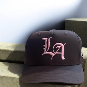 Puede incluir: Gorra de béisbol gris oscuro con las letras "LA" bordadas en rosa claro, con un tipo de letra estilizado. La gorra tiene una visera curva y una corona estructurada. El fondo es gris claro.