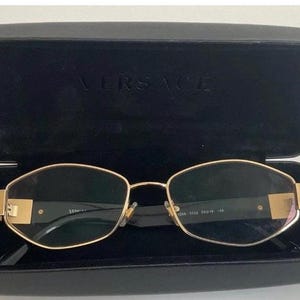 May include: Gold-framed spectacles with a rectangular shape, inside an open black case. The case displays the brand name "VERSACE". The glasses have dark lenses and a gold frame.