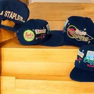Peut inclure: Quatre casquettes de baseball bleu marine ornées de patchs brodés colorés et de texte. Les motifs incluent LA STAPLES, Earl's, Pann's, Randy's Donuts et Dulan's Soul Food. Chaque casquette possède une calotte structurée et une visière plate.