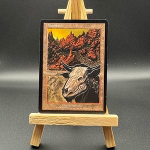 May include: A collectible card featuring a mountain landscape with a skull of a bull. The card is displayed on a small wooden easel. The card's text reads "Mountain" and "Land".